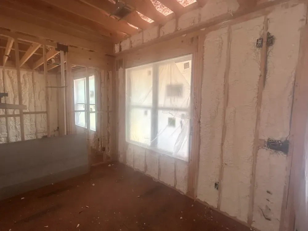 Interior of a room under construction, with spray foam insulation visible on walls and ceiling, window frames.