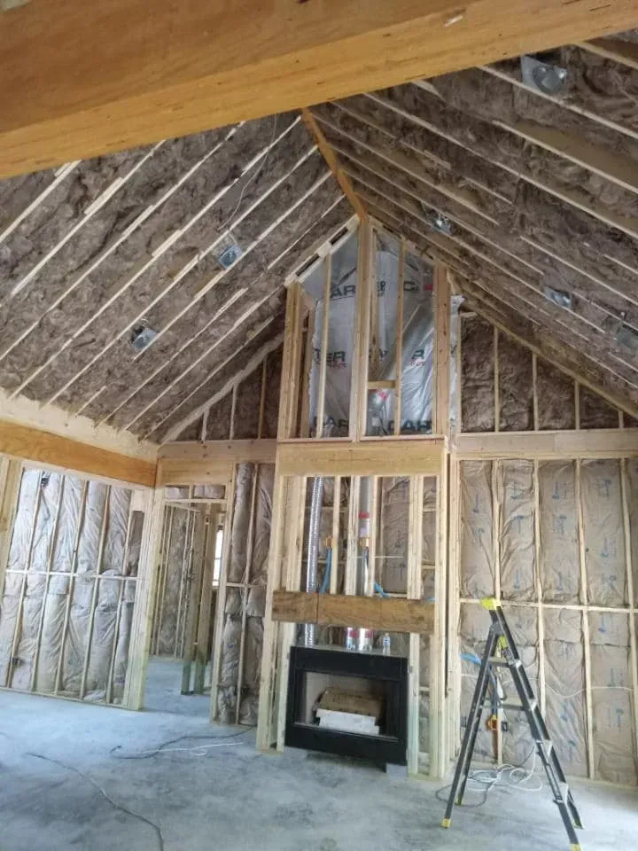 Interior of a house under construction; exposed wooden frame, insulation, fireplace, and a ladder.