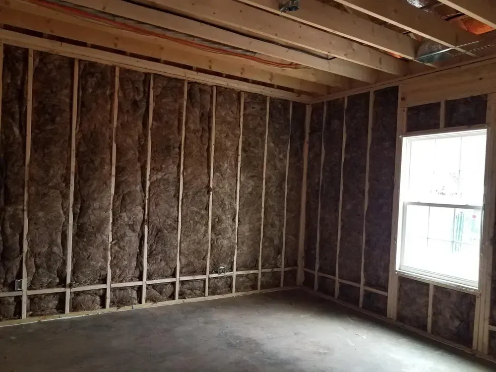 Interior of a room under construction; insulation installed in walls, studs visible, concrete floor, window.
