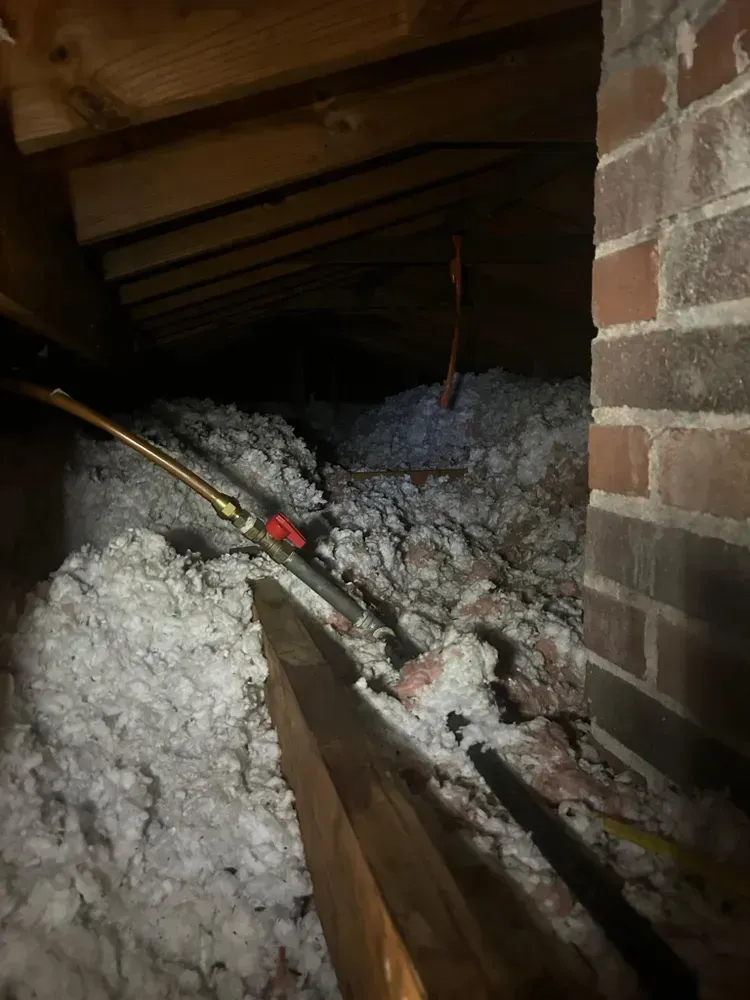 Attic interior with insulation, pipes, and brick wall. Dark with low ceiling.
