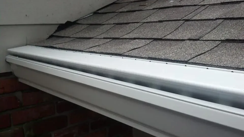 White rain gutter attached to a roof with brown shingles, resting on a brick wall.