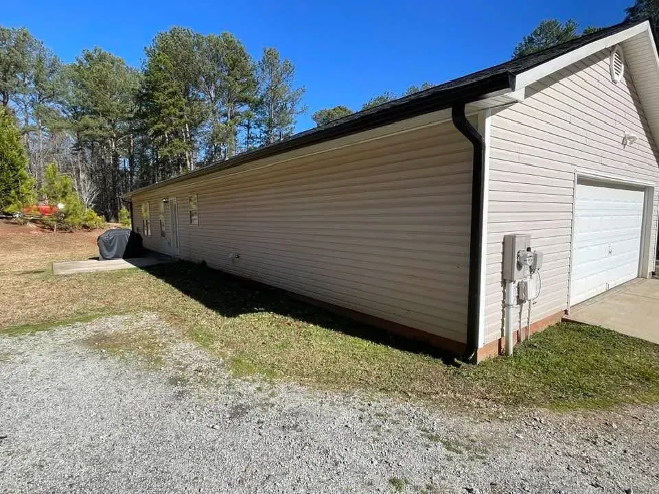 Garage with tan siding and white garage door, set on a gravel driveway, with trees in background.