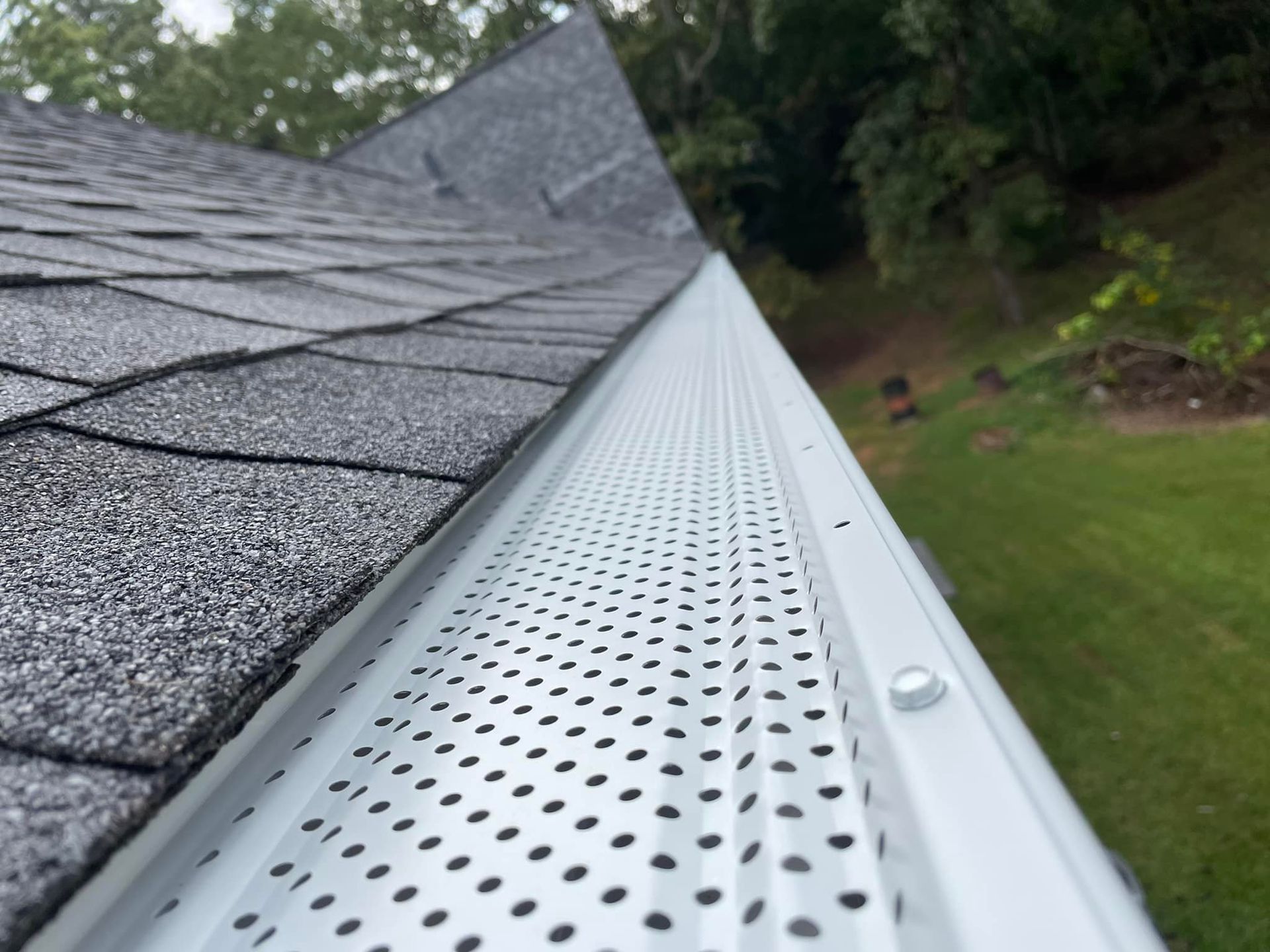 White gutter with perforated cover attached to a gray shingle roof, green grass in the background.