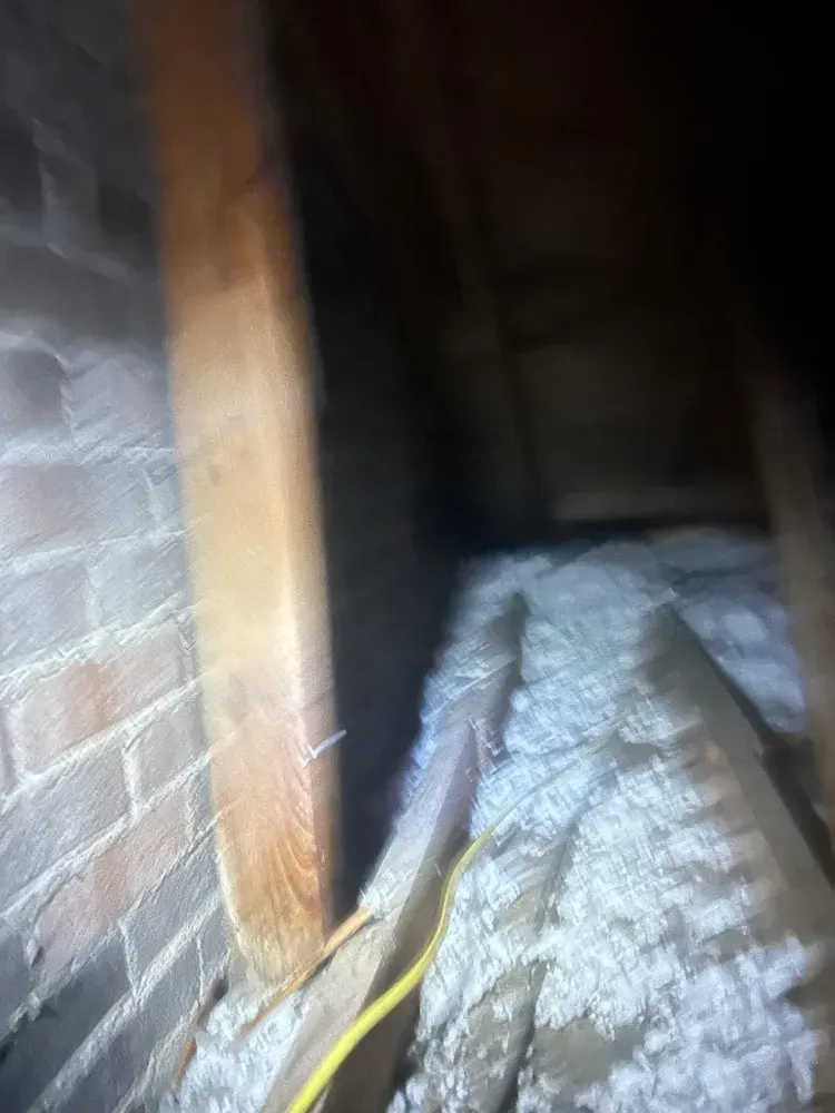 Attic interior view; wooden beam against brick wall, insulation visible, yellow wire. Dark, confined space.