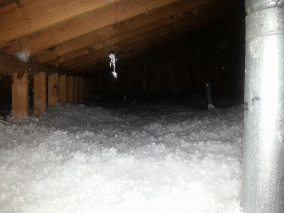 Attic with insulation, wooden beams, and a metal vent pipe. Dark interior, with visible light from the entrance.
