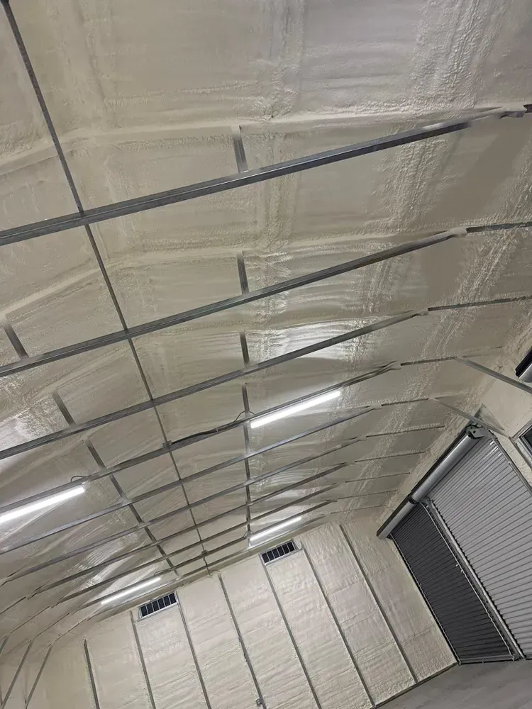 Interior view of a building's roof and walls, covered in white spray foam insulation, with metal beams.