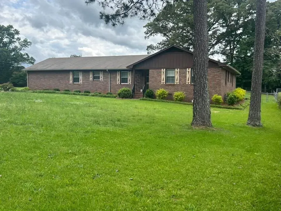 Brick ranch-style house with green lawn and trees under a cloudy sky.