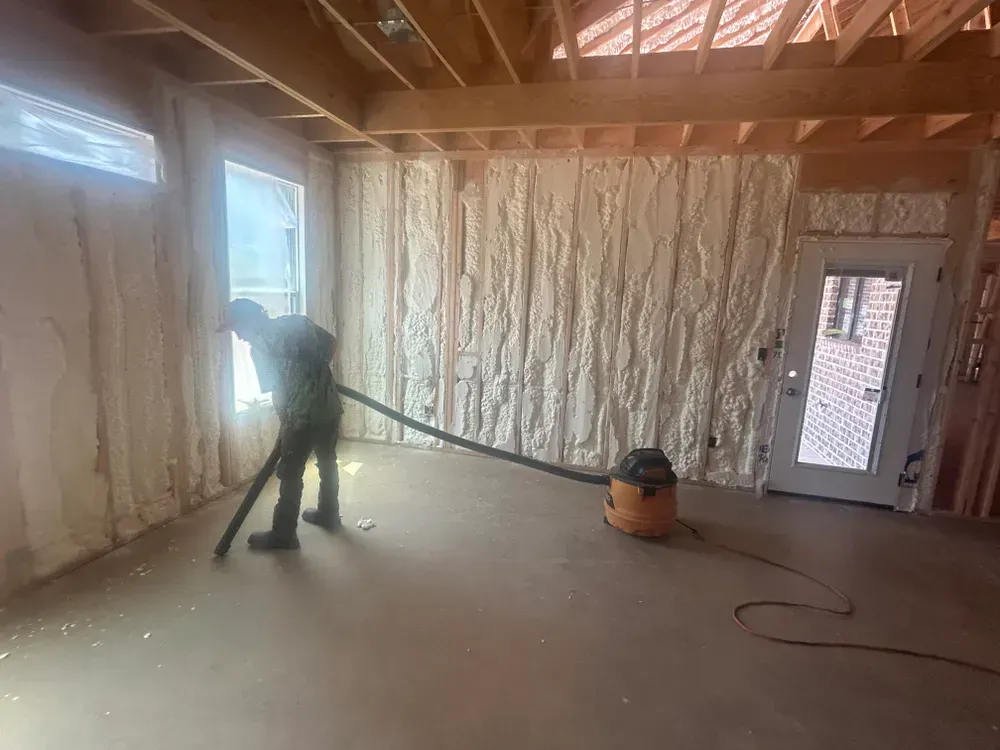 Man vacuums debris from a room with spray foam insulation. Window, door, and wooden framing visible.