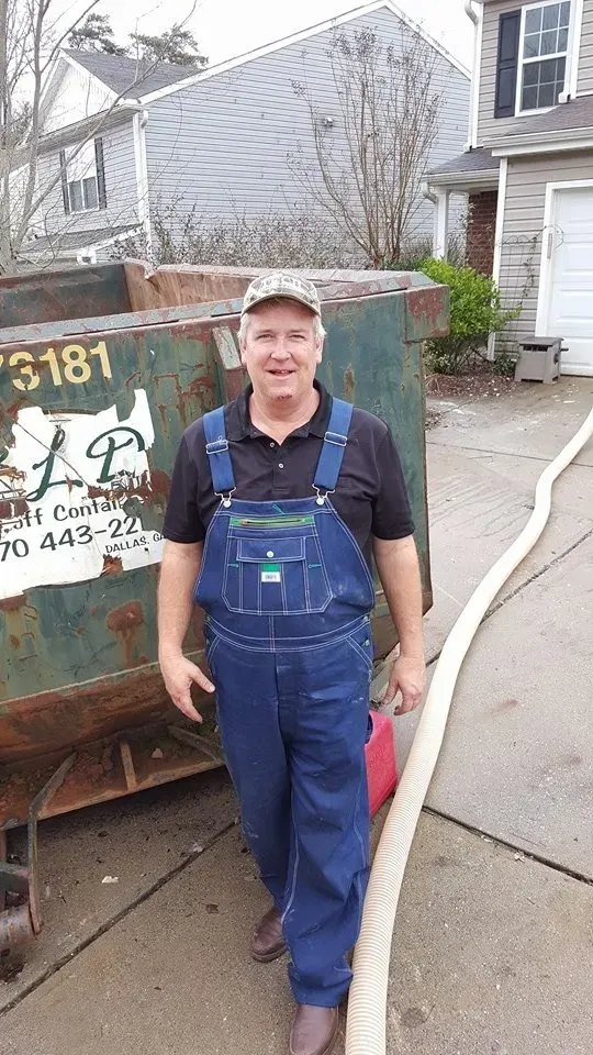 Man in blue overalls stands next to a dumpster and a hose. He wears a cap and smiles. Outdoors.