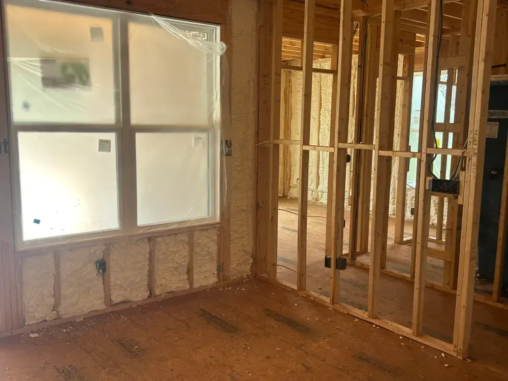 Interior of a room under construction, showing wooden studs, spray foam insulation, and a covered window.