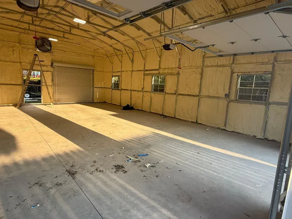 Interior view of a garage with spray foam insulation on the walls and ceiling. Sunlight streams in, illuminating the space.