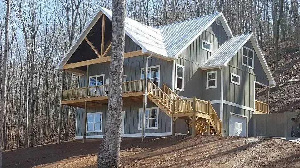 Two-story gray house with wood stairs, a metal roof, and a garage in a wooded area.