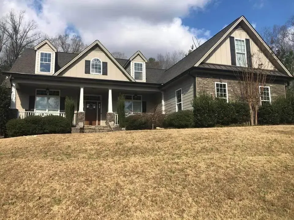 House with a stone and shingle exterior, front porch, and dormers on a grassy hill under a partly cloudy sky.