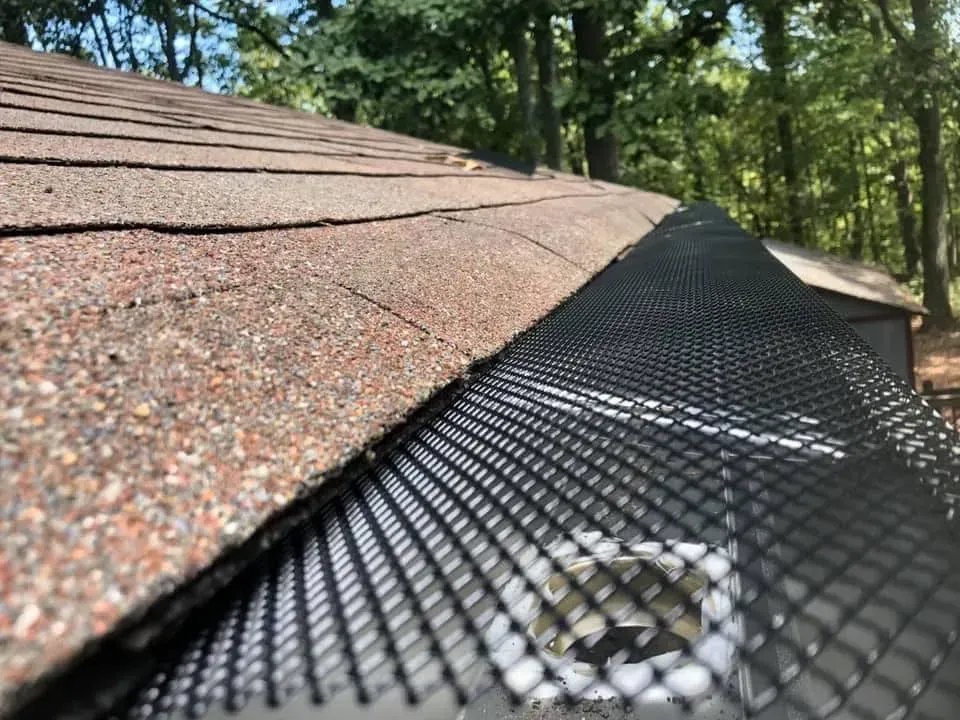 Close-up view of a roof with brown shingles, black gutter guard, and a forested background.
