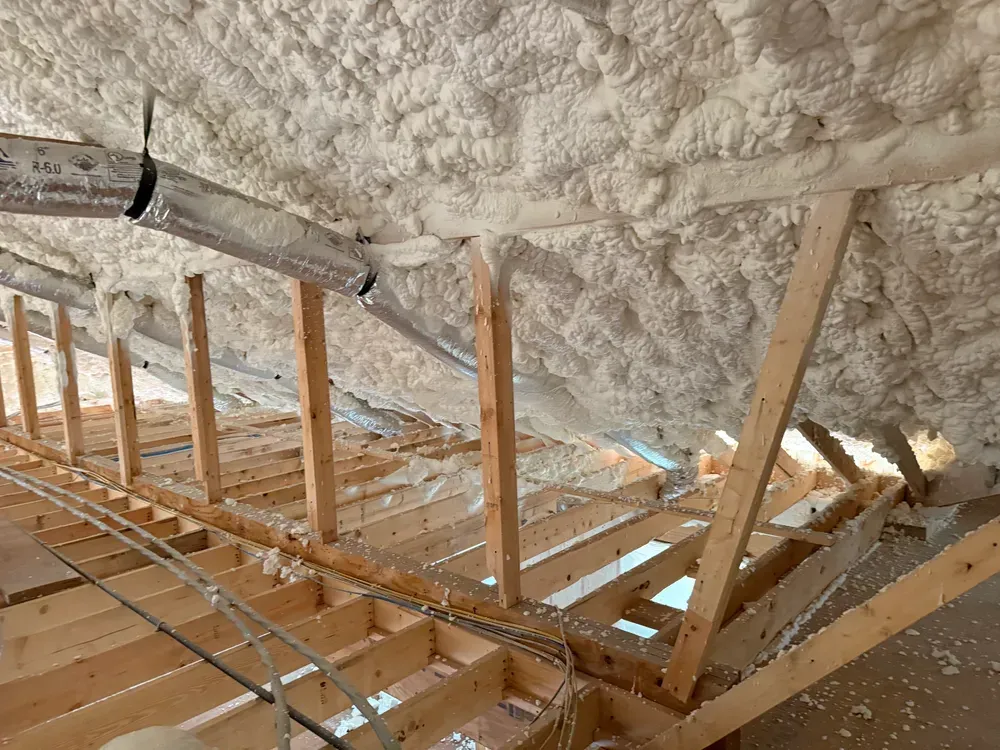 Wooden attic frame with spray foam insulation on the ceiling, pipes and wiring visible.