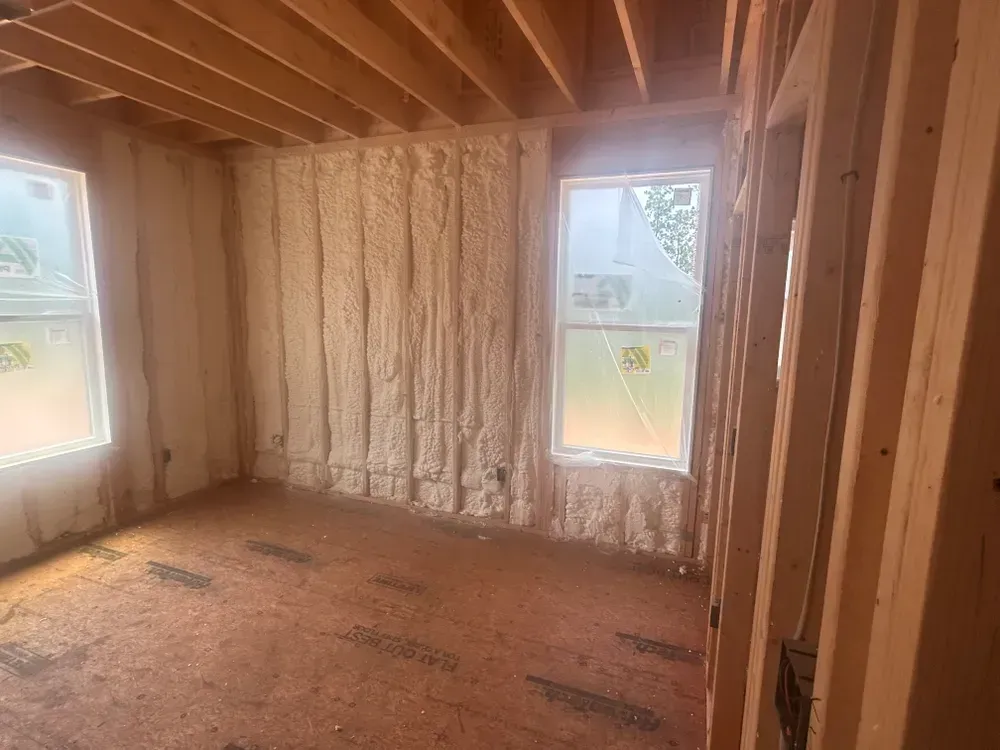 Interior view of a room with spray foam insulation on the walls and wooden framing; two windows.