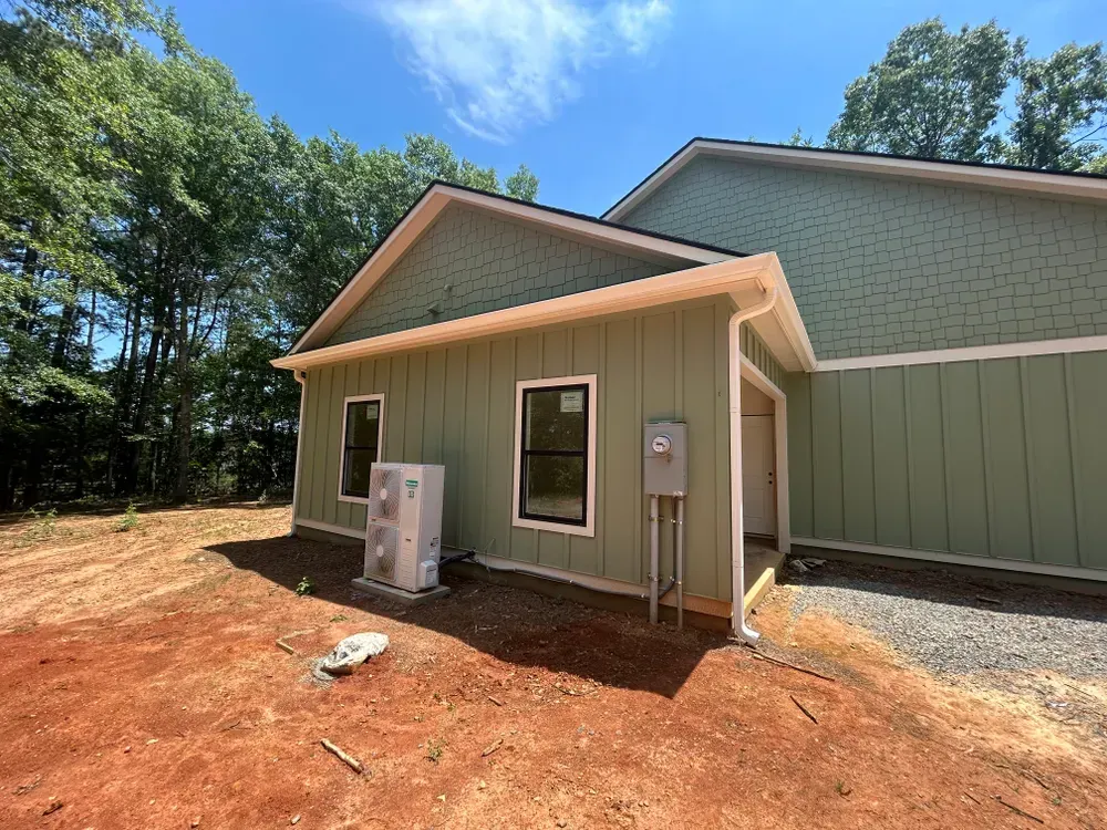 Green-sided building exterior, two windows, HVAC unit, utility box, and red dirt ground. Sunny day.