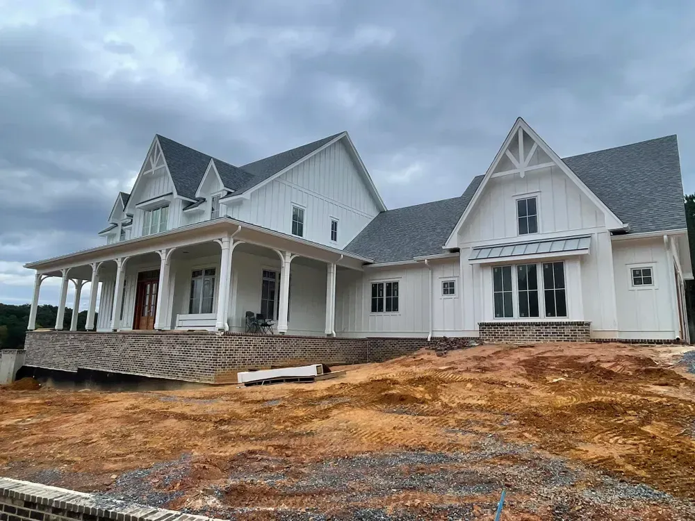 White farmhouse under construction on a dirt lot, with a porch and dark gray roof.