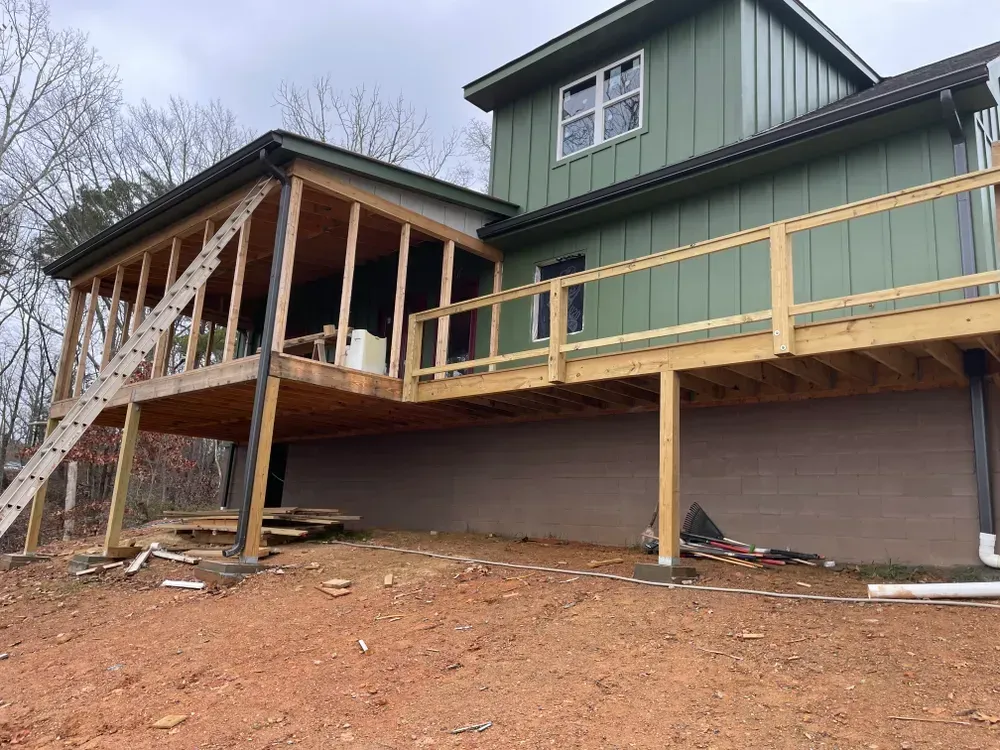 Exterior of a house with a green facade and wooden deck under construction; set on a brown, sloped yard.