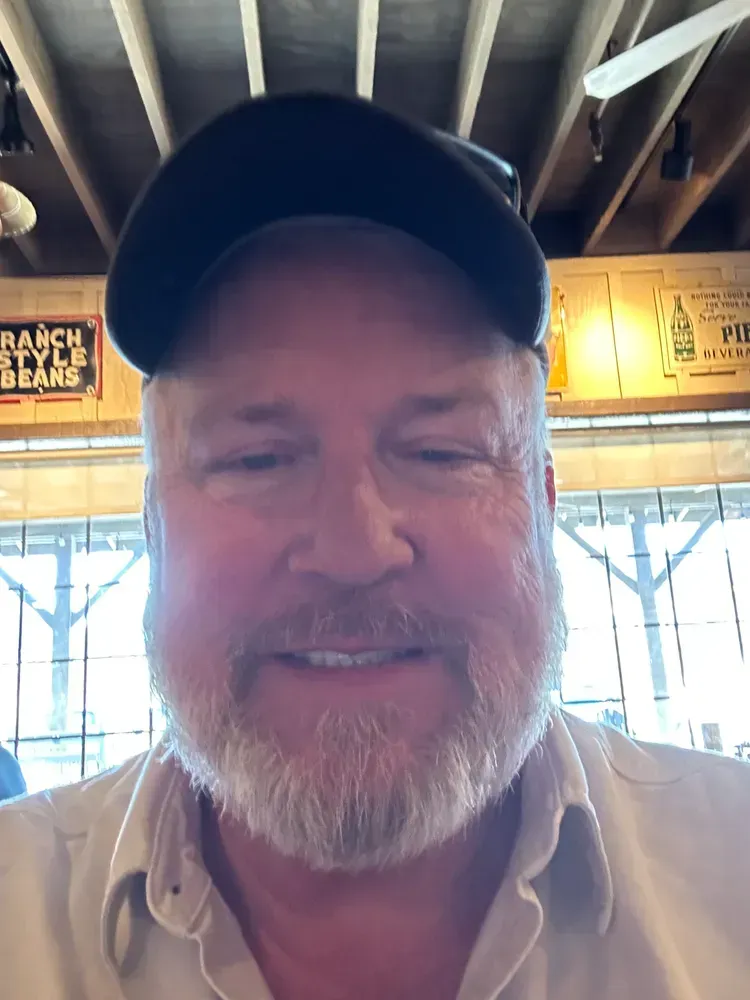 Man with a gray beard and black hat smiles at the camera, inside a restaurant.