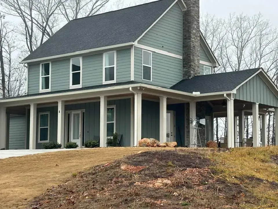 Two-story teal house with wrap-around porch and stone chimney, set on a slightly elevated, bare dirt lot.