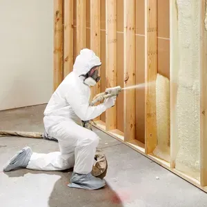 Person in protective suit spraying foam insulation into a wall frame in an indoor setting.