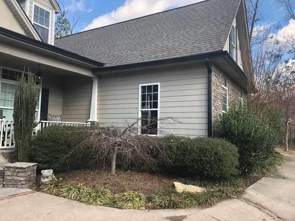 House exterior with tan siding, dark roof, bushes, and a small front yard.