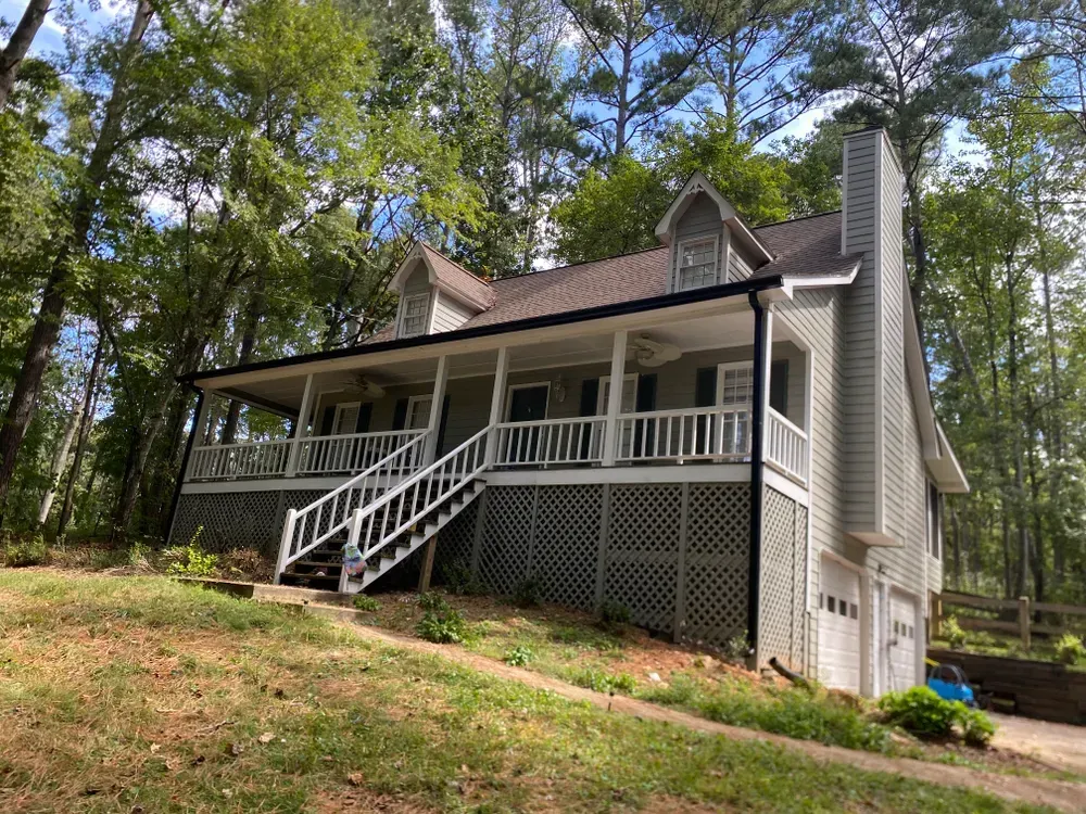 Two-story light gray house with a porch and garage, set on a hillside amid trees.