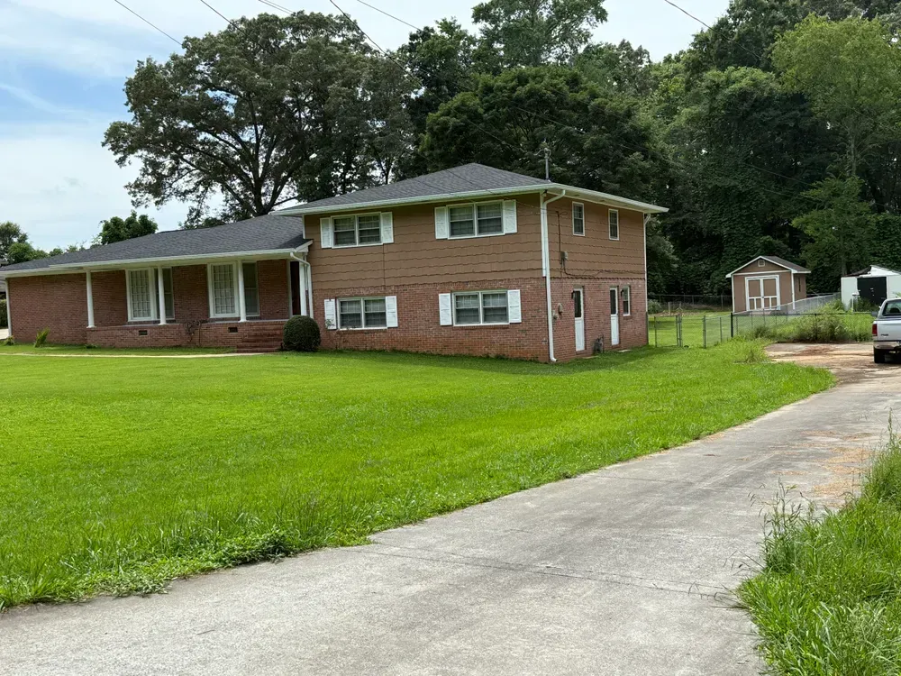 Two-story brick house with green lawn, driveway, and small shed; trees in the background under a blue sky.