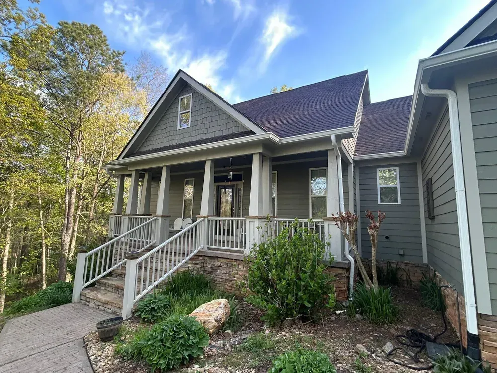 Green house with porch and steps, surrounded by trees and greenery under a blue sky.