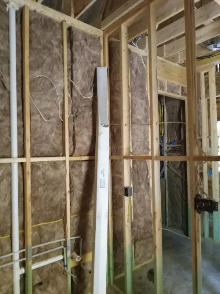 Interior of a building under construction. Wood framing with insulation in walls. Plumbing and electrical wiring visible.