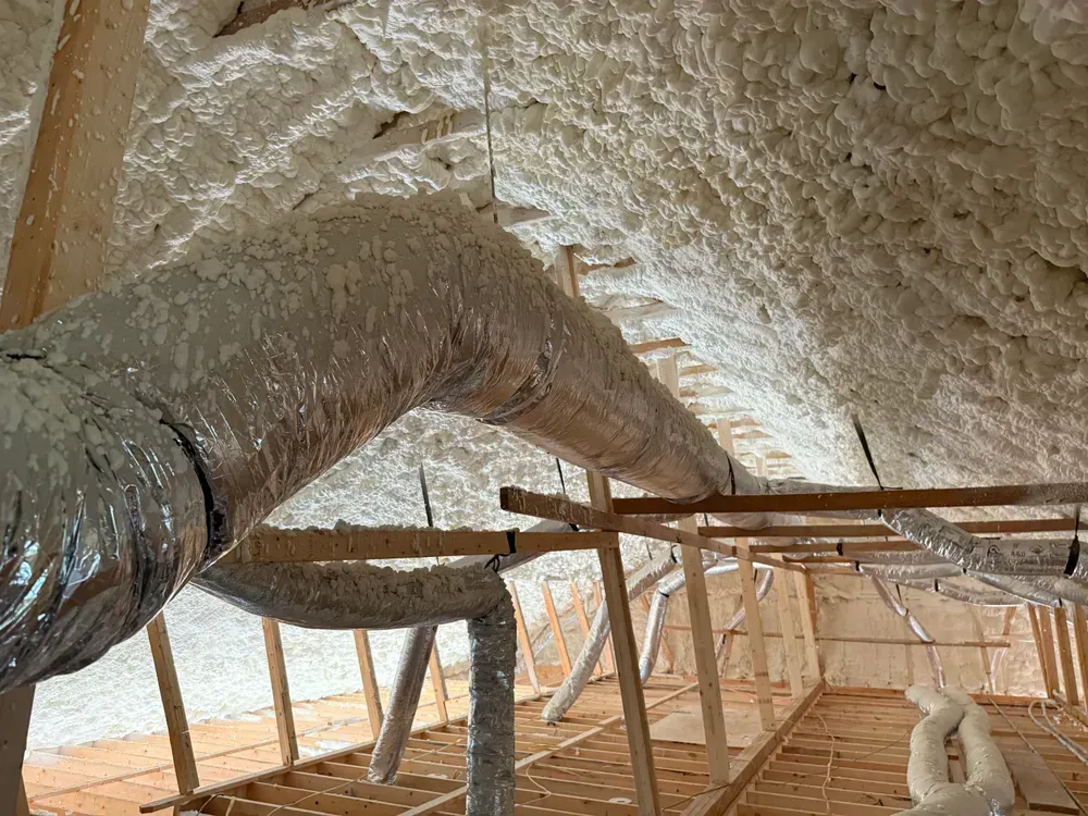 Attic interior with spray foam insulation surrounding ductwork and wooden framing.