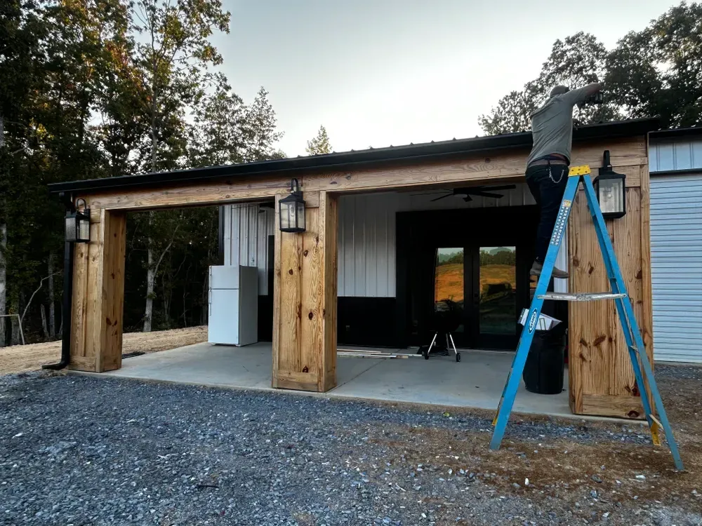 Person on ladder working on a building's exterior. The building has wood beams, metal roof, and hanging lights.
