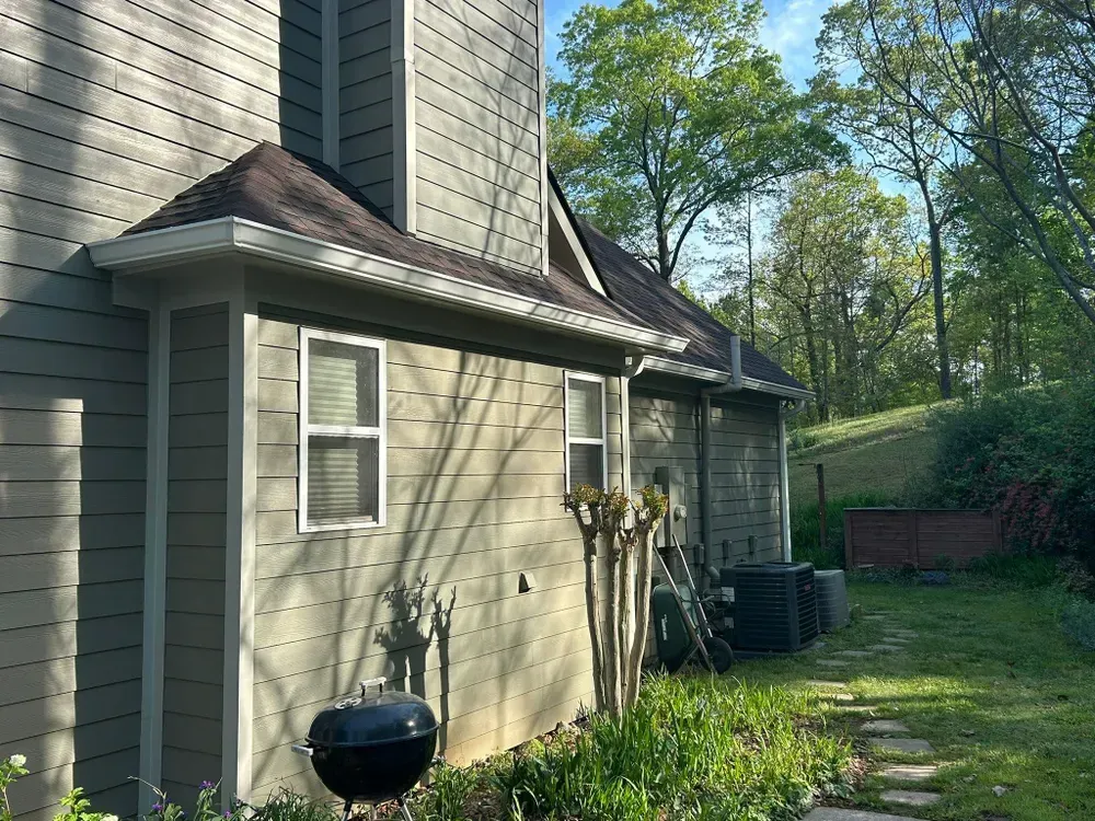 Side view of a house with olive-green siding, a dark roof, and a black grill in a grassy yard.