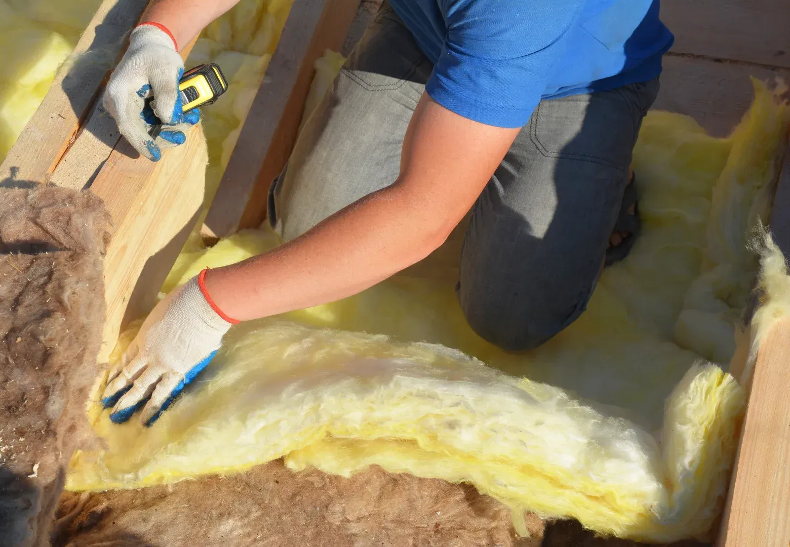 Person installing yellow insulation between wooden beams, wearing gloves.