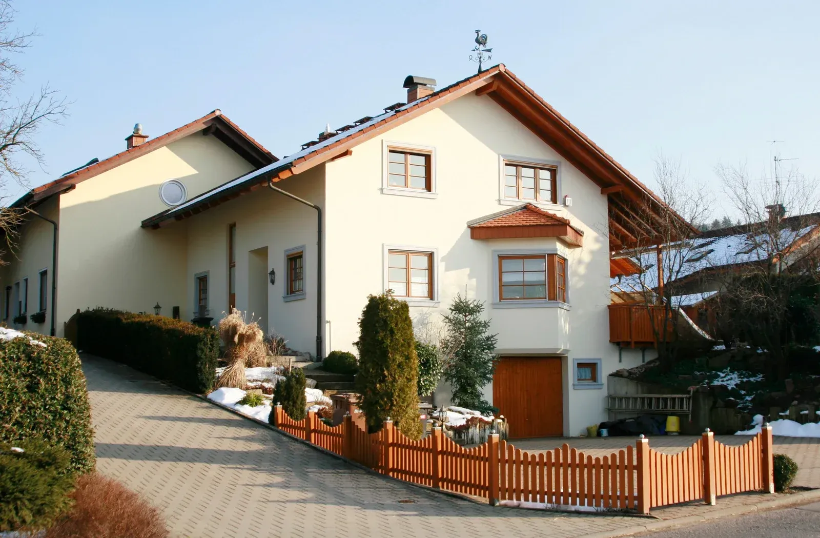 Two-story beige house with brown roof and trim, wooden fence, driveway, and snow patches.
