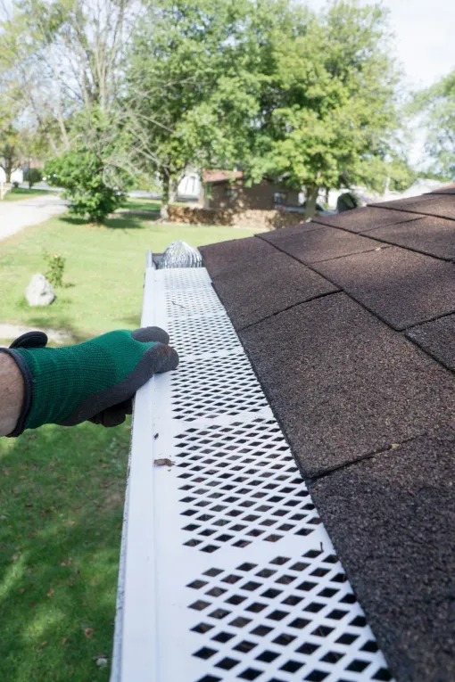 Gloved hand holding white gutter guard on a rooftop; green lawn and trees in the background.