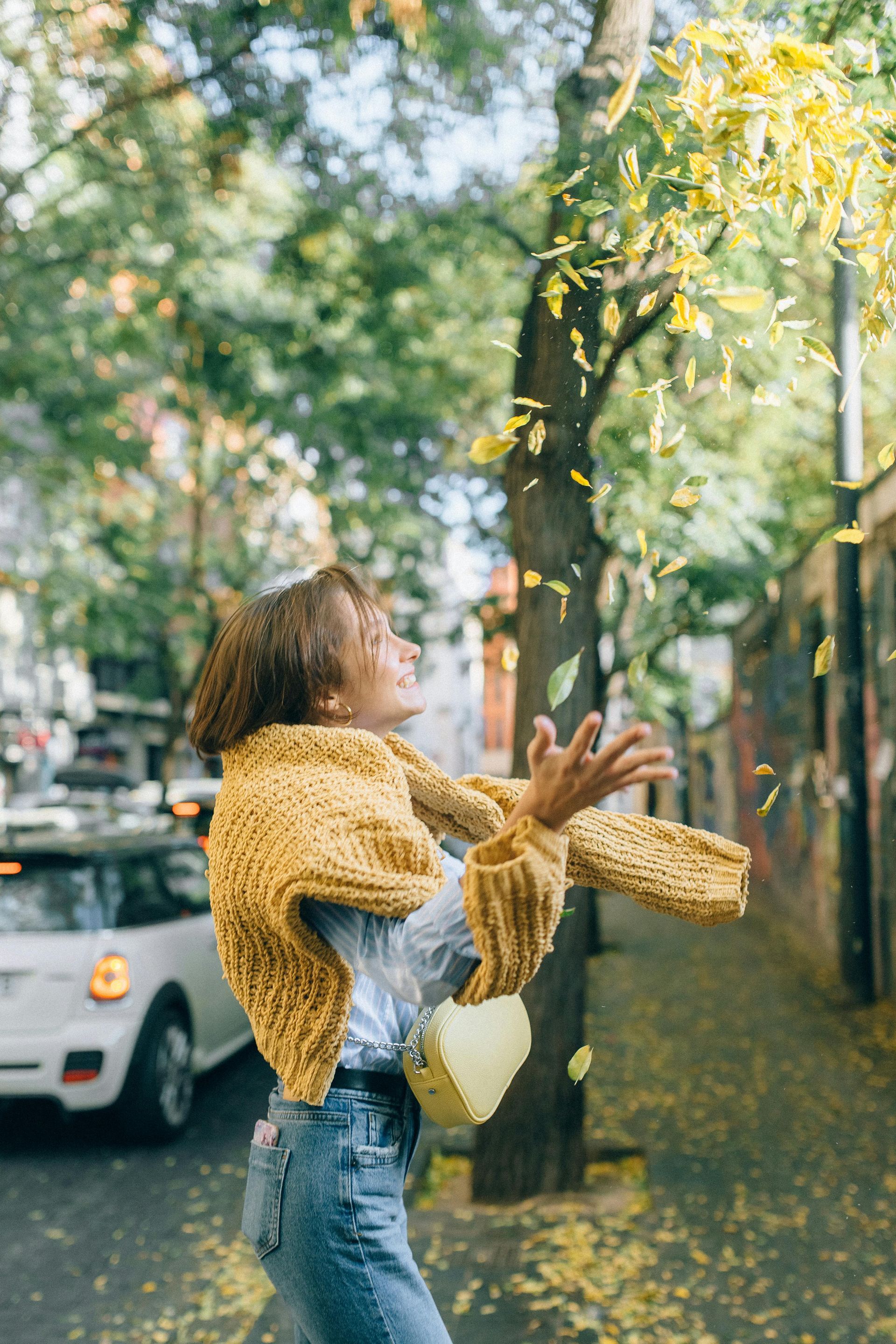 A woman in a yellow sweater is throwing leaves in the air.