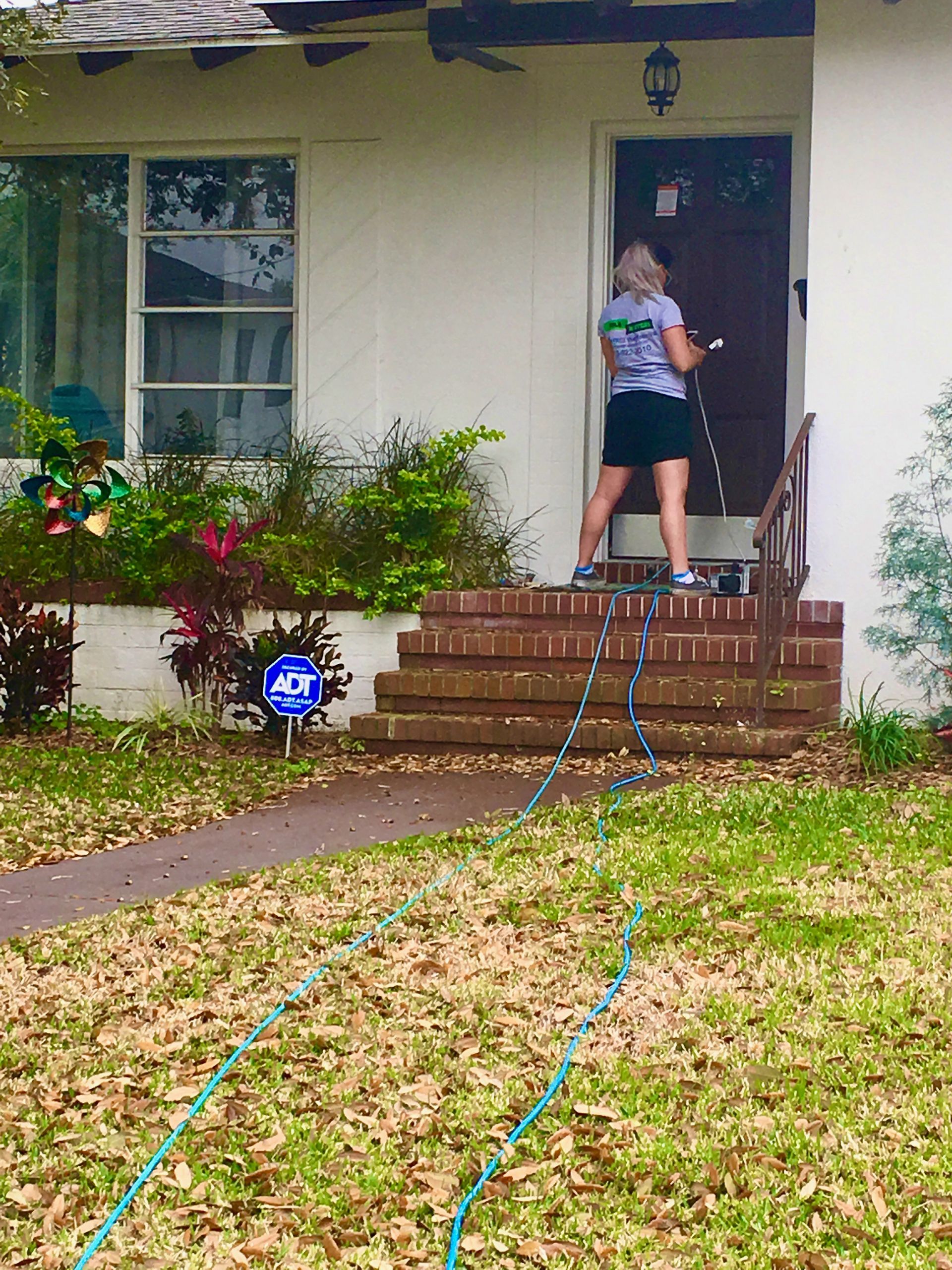 Woman on porch pulling green hoses, facing a white house with brick steps and a window.