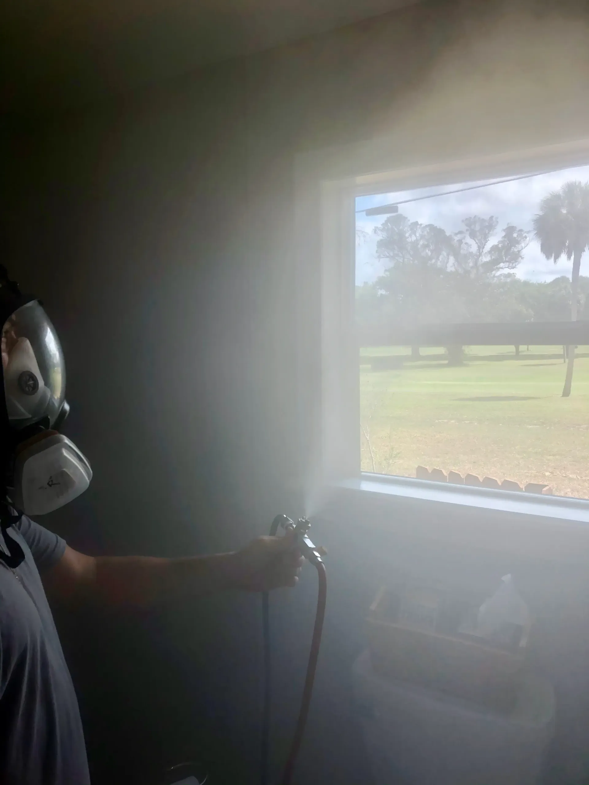 Person in respirator sprays paint indoors near a window, filling the room with mist.