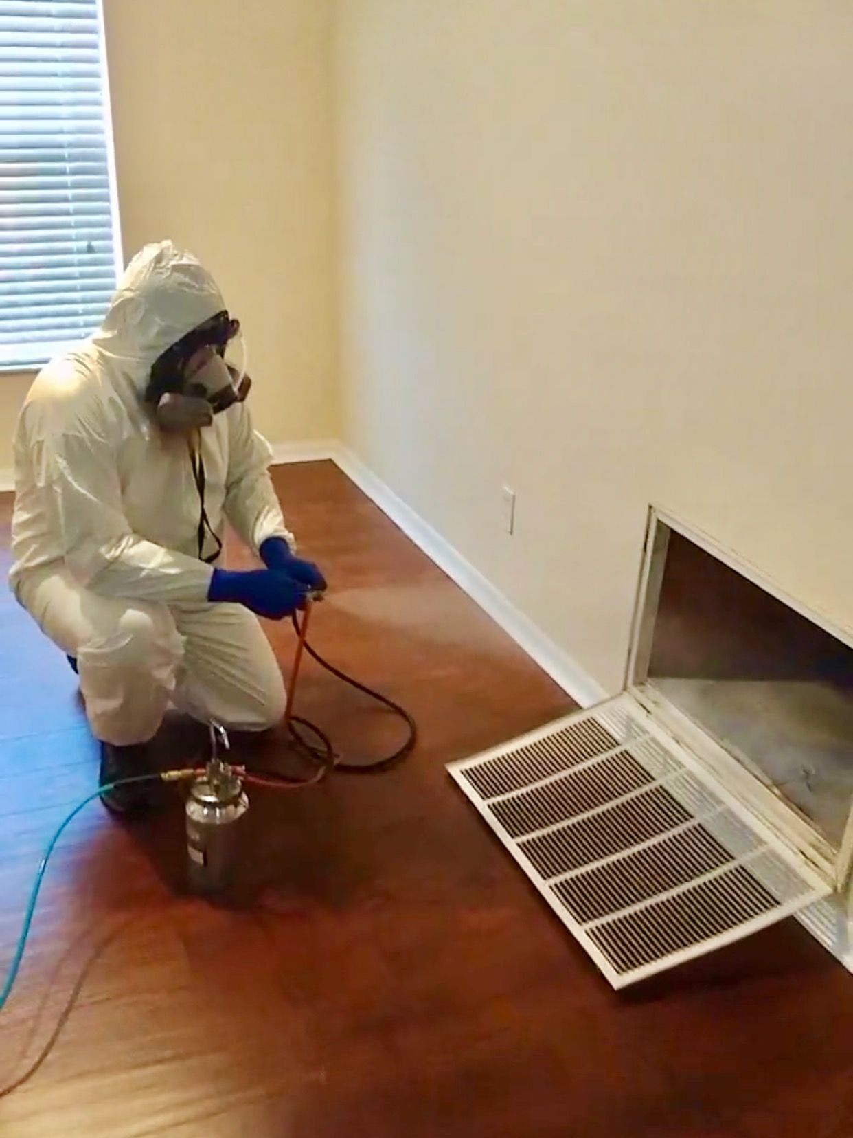 Person in hazmat suit sprays into an air vent in a room with hardwood floors.