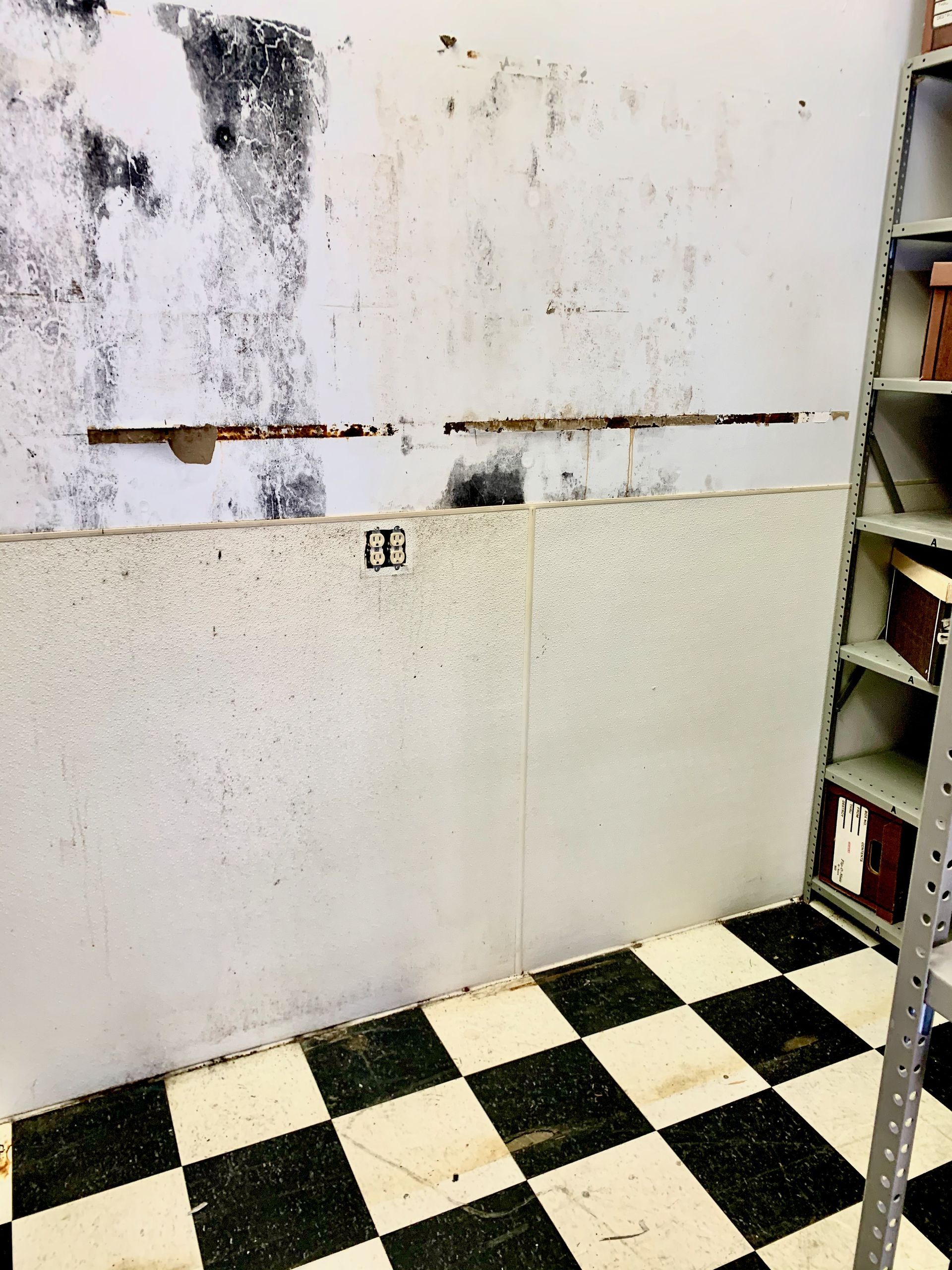 Wall with water damage, above checkered floor, next to metal shelving.