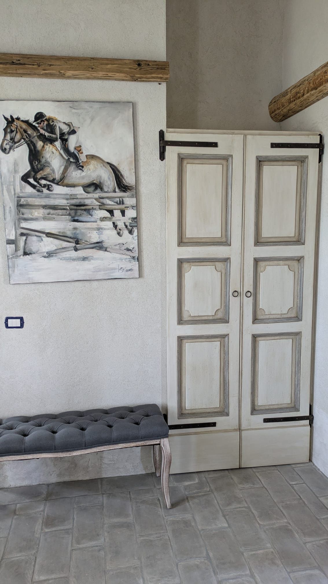 Dipinto equestre sopra una panca imbottita. Porte bianche con ferramenta nera accanto. Ambiente rustico.