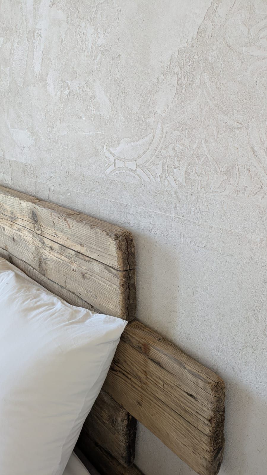 Testiera del letto in legno rustico con cuscino bianco, contro una parete chiara e strutturata.