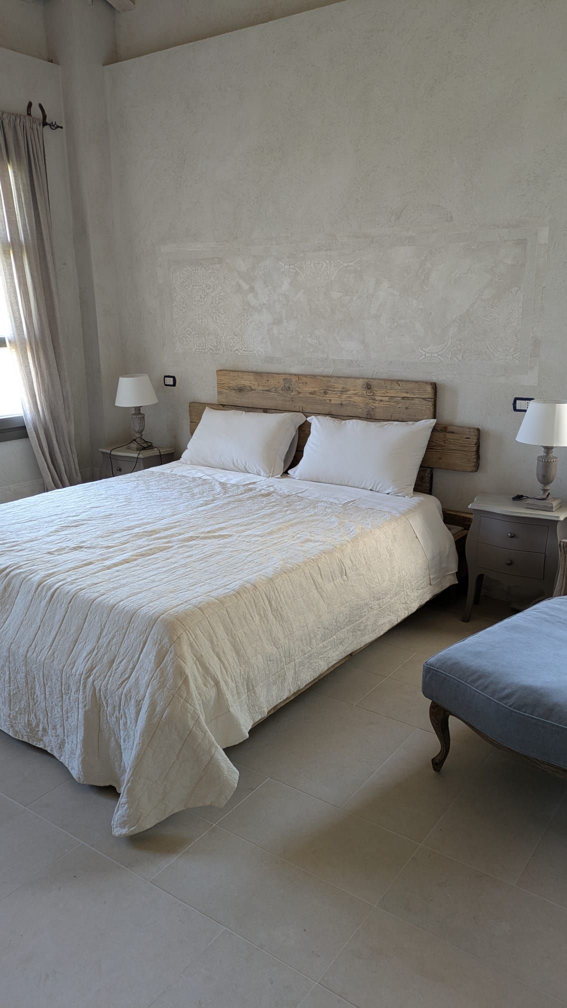 Camera da letto con letto, comodini, lampade e un poggiapiedi blu. Pareti e pavimento bianco crema.