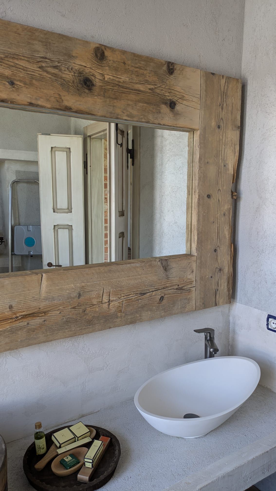 Bagno con uno specchio con cornice in legno sopra un lavandino bianco ovale su un piano di lavoro in cemento.