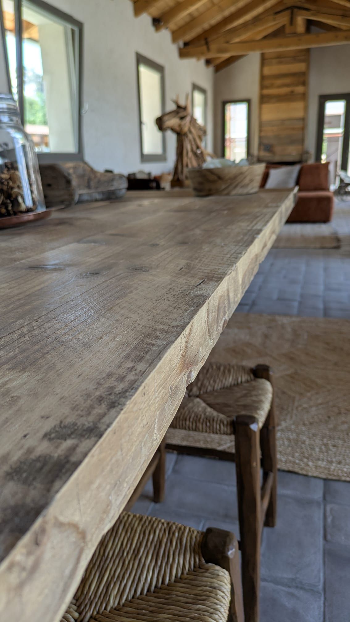Tavolo rustico in legno con sedie intrecciate, in una stanza con grandi finestre e una scultura decorativa a forma di cavallo.