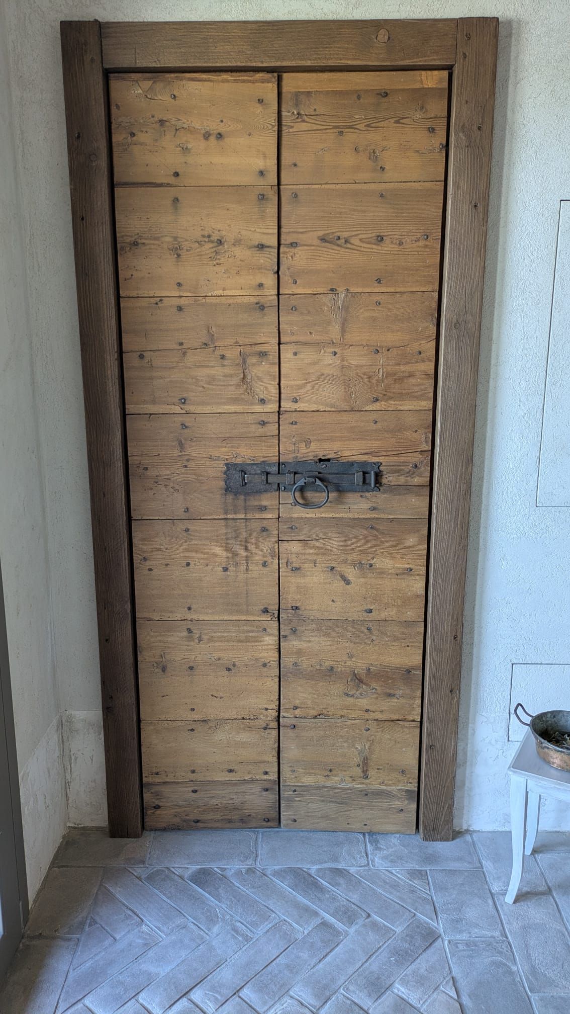 Porta doppia in legno con ferramenta rustica, inserita in una cornice in legno testurizzato e appoggiata su piastrelle a spina di pesce.