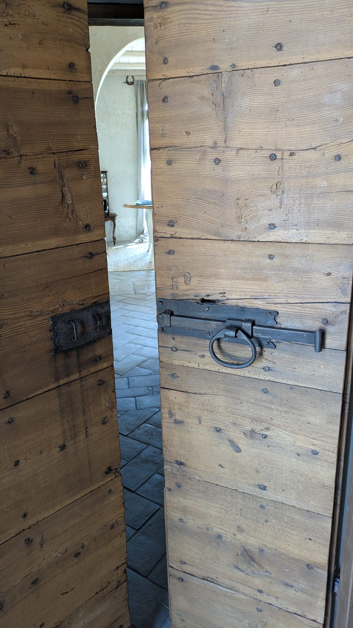 Doppie porte in legno, una delle quali leggermente socchiusa, che lasciano intravedere una stanza interna con pavimento in pietra. Ferramenta in metallo pesante.
