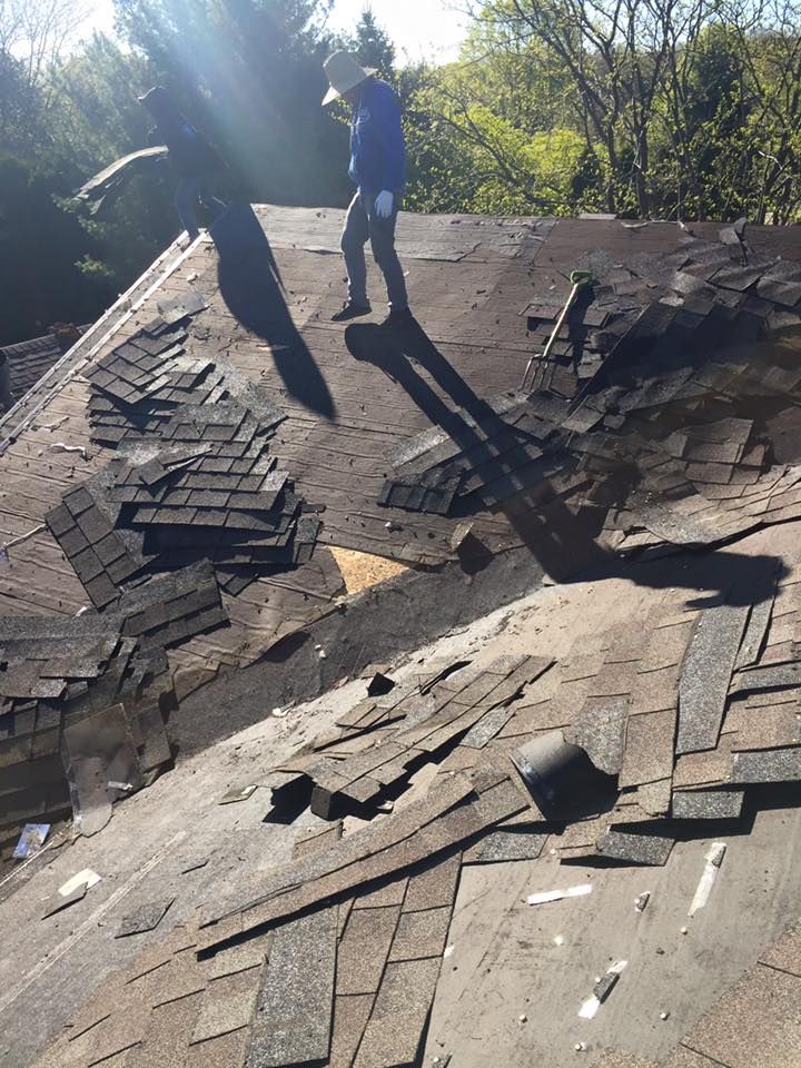 A person wearing a hat and blue shirt stands on a sloped roof amidst torn-up asphalt shingles during a roofing project.
